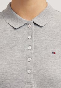 Grå polo skjorta gjord av strukturerad tyg, med klassisk krage, fem knappar och en liten broderad logotyp i röd, vit och blå.