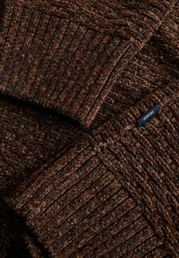 Dettaglio di un maglione lavorato a maglia marrone che mostra la texture a coste, un tessuto morbido e un piccolo etichetta del brand. Diverse sfumature di marrone creano profondità.