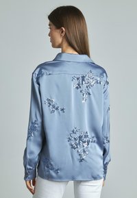 Camicia di raso azzurro chiaro con ricami floreali in paillettes, maniche lunghe e colletto. Texture liscia e vestibilità rilassata, cuciture dettagliate visibili.
