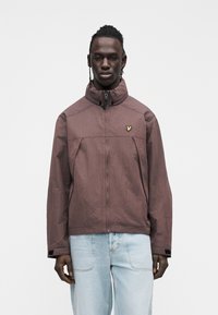 Giacca marrone con cerniera, collo alto, tessuto testurizzato e un piccolo logo dorato sul petto. Abbinata a jeans azzurri.