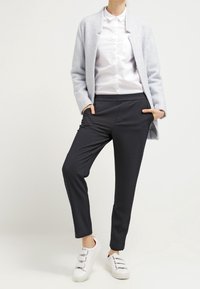 Cardigan grigio chiaro, camicia bianca con bottoni, pantaloni scuri slim e sneakers bianche. I tessuti sono morbidi con minima texture; vestibilità moderna e su misura.