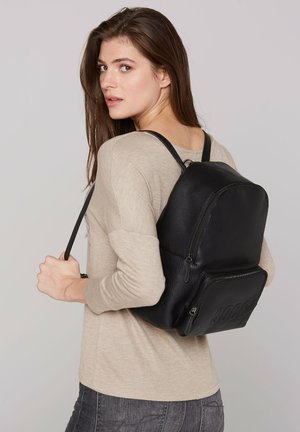 Schwarzer Leder-Rucksack mit glatter Textur, mit Reißverschlussfächern und erhabenem Logo. Über einer Schulter getragen, was sein kompaktes Design zur Geltung bringt.