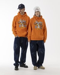 Orange hoodies with graphic prints, paired with dark blue baggy jeans featuring a repeated pattern. The model wears a white beanie and a blue cap.