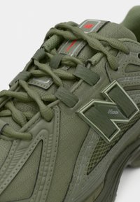 Baskets vert olive avec un tissu texturé, des lacets coordonnés et un logo "N" bien visible sur le côté. Accents en mesh pour la respirabilité.