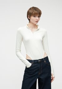 Λευκό κορμάκι με μακριά μανίκια, φερμουάρ στον γιακά και ανοιχτές πλευρές, σε συνδυασμό με σκούρο μπλε παντελόνι με φαρδιά πόδια, το οποίο διαθέτει μπροστινές τσέπες και αντίθετη ραφή.