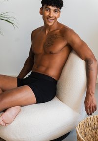 Schwarze Boxershorts, helle Hautfarbe, lockiges Haar, sitzend auf einem weißen, strukturierten, geschwungenen Stuhl. In der Nähe ein geflochtener Korb mit unregelmäßigem Rand.