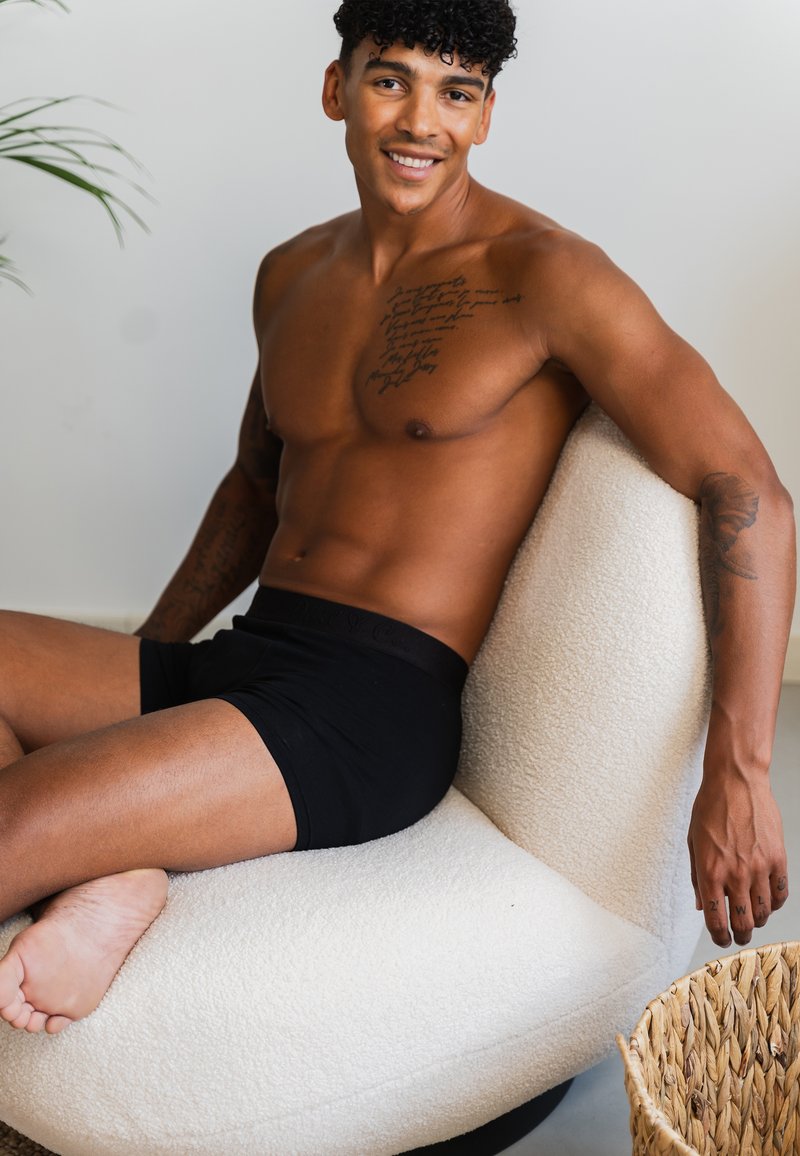 Schwarze Boxershorts, helle Hautfarbe, lockiges Haar, sitzend auf einem weißen, strukturierten, geschwungenen Stuhl. In der Nähe ein geflochtener Korb mit unregelmäßigem Rand.
