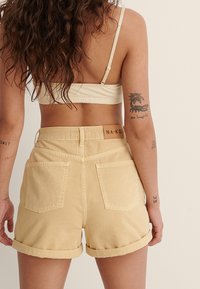 Pantalones cortos de mezclilla amarillos con dobladillos enrollados, que cuentan con dos bolsillos traseros y una etiqueta de la marca en la cinturilla. Textura suave y diseño ajustado.