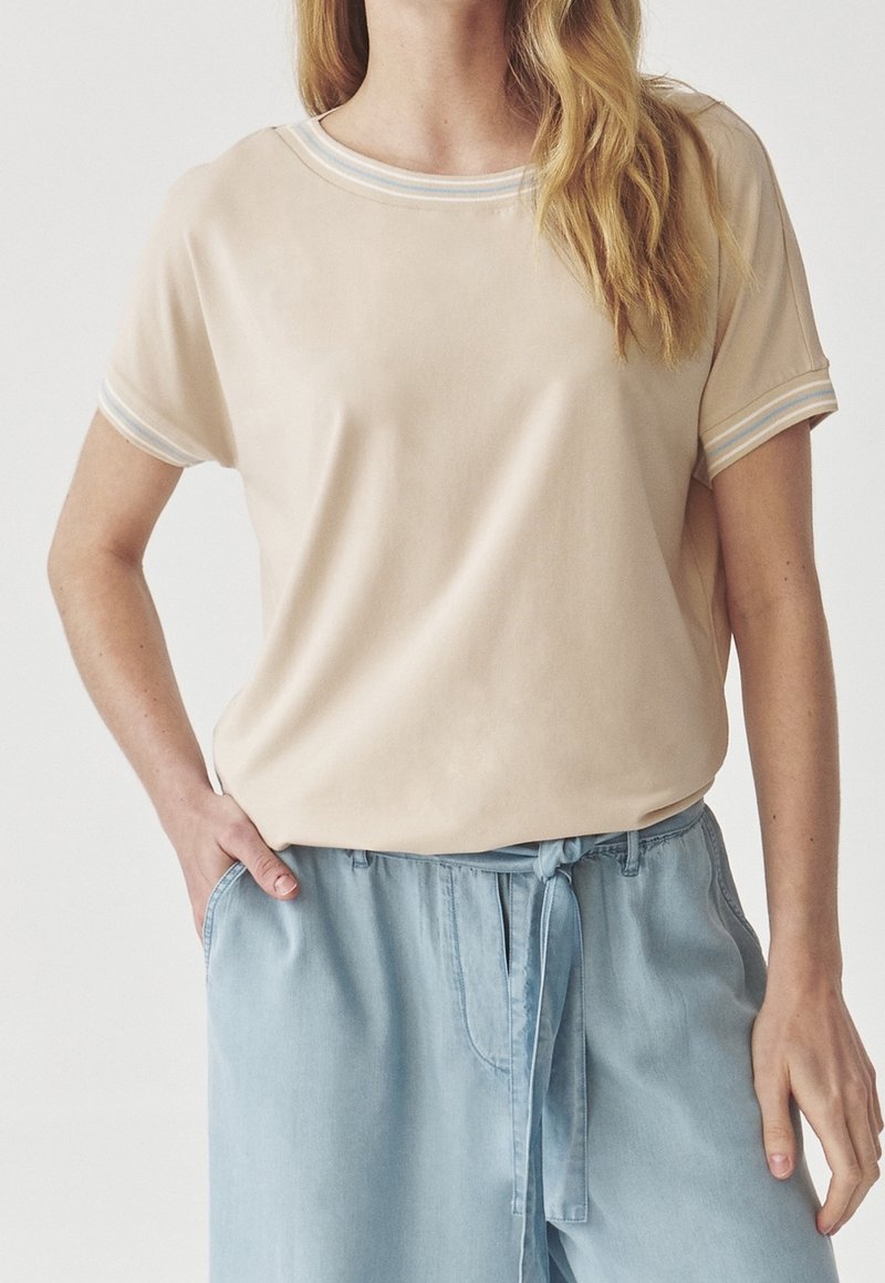Beige t-shirt med korte ærmer og rund halshull, med lys blå og hvid stribet kant langs kraven og ærmerne.