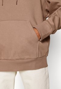 Sweat à capuche en coton marron avec poignets et ourlet côtelés, doté d'une poche avant. Associé à un pantalon de couleur claire. Texture douce, design décontracté.