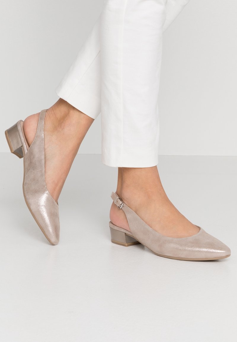 Pieds portant des chaussures slingback à talon bloc bas beige clair, associés à un pantalon blanc court sur un fond uni.