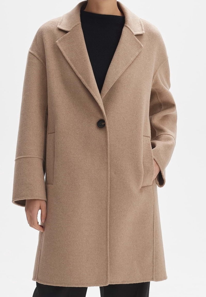 Manteau en laine beige avec revers, fermeture à un bouton, et poches avant. Présente une coupe décontractée et un tissu texturé.