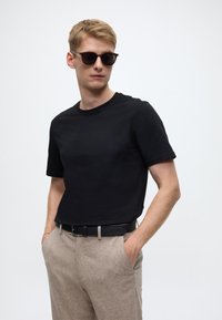 Schwarzes kurzärmeliges T-Shirt kombiniert mit hellbrauner Hose mit strukturierter Oberfläche, getragen mit schwarzem Gürtel und Sonnenbrille.