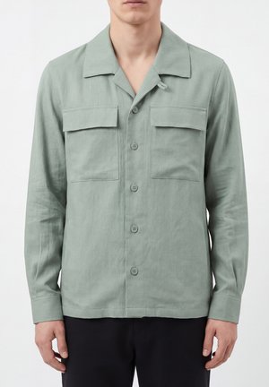 Hombre con camisa verde salvia abotonada con dos bolsillos en el pecho y pantalones negros, de pie sobre un fondo blanco liso.