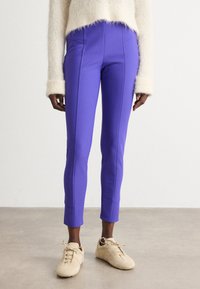 Paarse, slim-fit broek met contrasterende naden, cropped lengte en soepele textuur, gecombineerd met beige sneakers op een neutrale vloer.