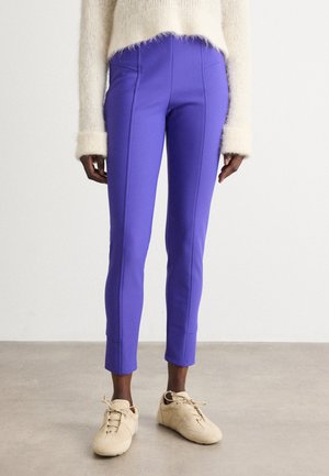 Purple, slim-fit pants with contrasting seams, cropped length, and smooth texture, paired with beige sneakers on a neutral floor.