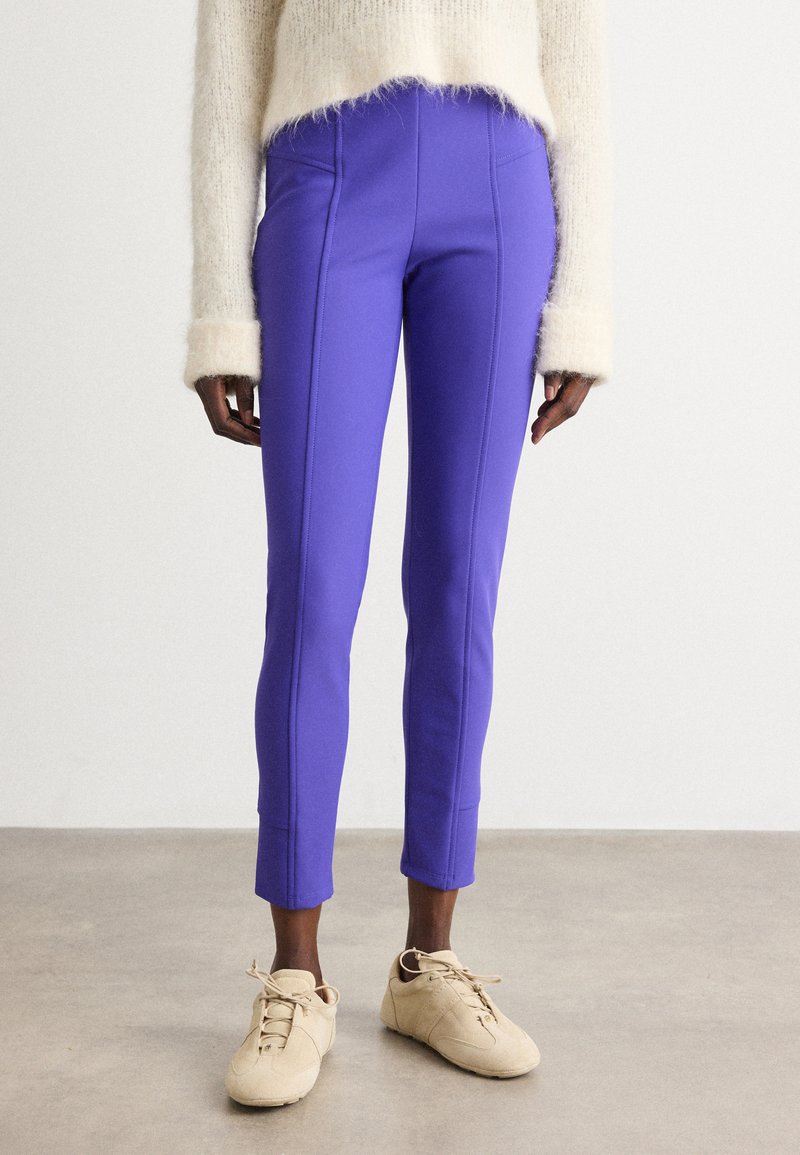 Paarse, slim-fit broek met contrasterende naden, cropped lengte en soepele textuur, gecombineerd met beige sneakers op een neutrale vloer.