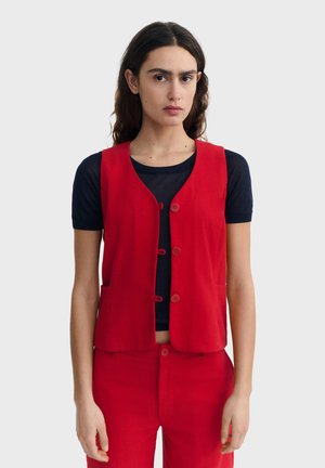 Femme aux cheveux foncés portant un gilet rouge sur un t-shirt noir à manches courtes et un pantalon rouge, debout devant un fond clair uni.