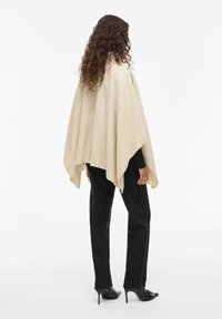 Beige oversized poncho lavet af tekstureret stof, med en trekantet form og rundede kanter. Båret med sorte lige ben-bukser.