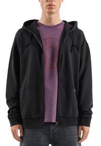 Czarna bluza z kapturem na zamek błyskawiczny, z regulowanym sznurkiem, wykonana z mieszanki bawełny, z przednimi kieszeniami, noszona na fioletowej koszulce z okrągłym nadrukiem.