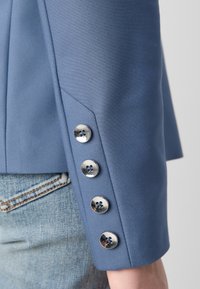 Gros plan de la manche d'un blazer bleu avec quatre boutons métalliques brillants au-dessus de la main d'une personne et un jean en denim bleu.
