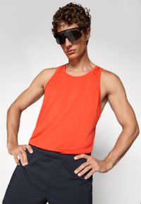 Helles oranges Tanktop aus strukturierter Stoff, mit weiten Armausschnitten und rundem Ausschnitt, kombiniert mit dunklen Shorts und übergroßen schwarzen Sonnenbrillen.