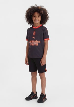 Enfant souriant aux cheveux bouclés portant un maillot de football noir et orange de l’AC Milan, un short noir et des baskets Puma noires, debout sur fond blanc.