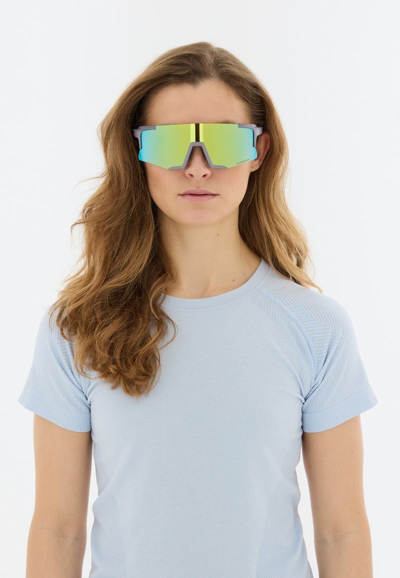 Donna con capelli lunghi e mossi, che indossa una camicia a maniche corte azzurro chiaro e grandi occhiali da sole sportivi riflettenti, rivolta in avanti su sfondo bianco.