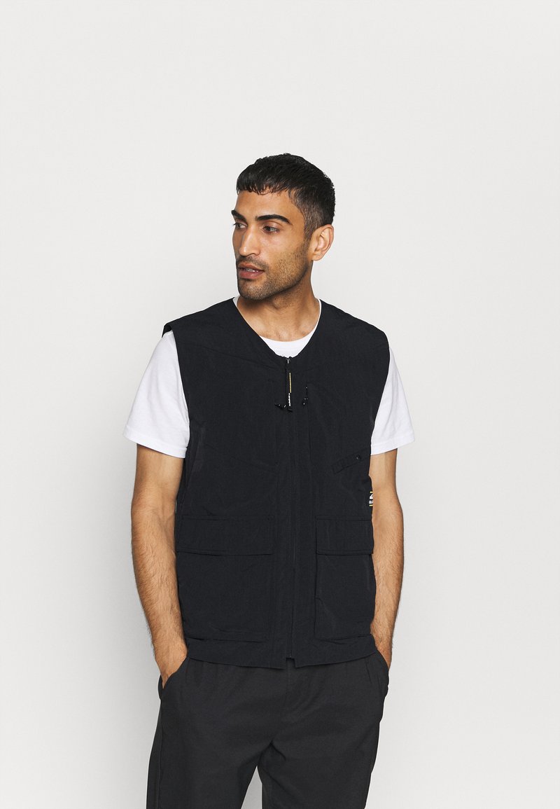 Colete sem mangas preto com fecho à frente e múltiplos bolsos, usado sobre uma t-shirt branca. Textura suave, design prático e estilo versátil.
