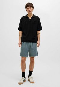 Polo shirt tricotat negru cu mâneci scurte, combinat cu pantaloni scurți din catifea ușoară de culoare albastru deschis și adidași albi cu accente verzi și roșii.
