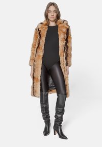 Faux fur coat in light brown with dark stripes, layered over a black bodysuit, paired with shiny black leggings and knee-high, heeled boots.