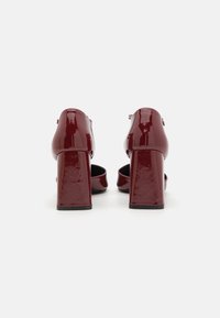 Scarpe con tacco alto in pelle verniciata bordeaux, con design a tacco quadrato e cinturino alla caviglia, caratterizzate da una finitura liscia e lucida.