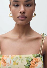 Bloemendress met een gele achtergrond, met oranje en groene patronen, dunne schouderbanden en gouden creolen. Soepele stoftextuur.