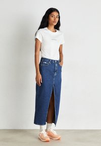 Džínová maxi sukně s předním rozparkem, v kombinaci s bílým tričkem a broskvovými teniskami. Oblečení se vyznačuje minimálními detaily a klasickým designem.