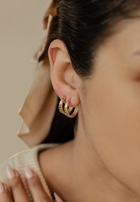 Boucles d'oreilles en or en forme de boucle, comprenant trois anneaux entrelacés, avec une finition polie et une texture subtile, portées sur l'oreille d'un modèle.