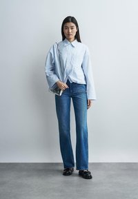 Fiatal nő kék csíkos ingben, kék farmerral, fekete cipőben, ezüst kézitáskát tartva, világosszürke fal előtt áll.