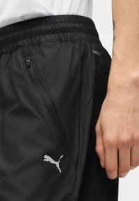 Μαύρο παντελόνι φόρμας Puma με φερμουάρ στην πλαϊνή τσέπη, ελαστική μέση και μερική όψη χεριού ατόμου και λευκού πουκάμισου.