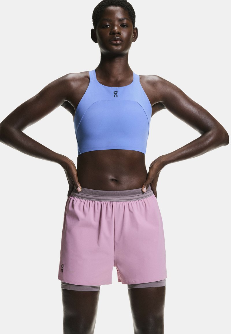 Sujetador deportivo azul claro con escote alto y tirantes, combinado con pantalones cortos rosas que tienen una cintura gris y detalle de logo.