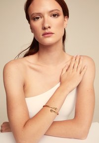 Bracelet en or sur un bras nu, avec un design lisse et courbé, doté de deux accents sphériques à chaque extrémité. Le teint de la peau est clair.