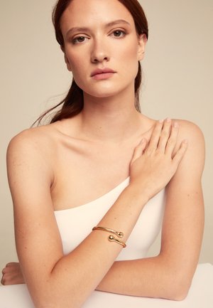 Bracelet en or sur un bras nu, avec un design lisse et courbé, doté de deux accents sphériques à chaque extrémité. Le teint de la peau est clair.