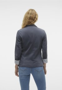 Šedý blazer s klopovým límcem a tříčtvrtečními rukávy, s prvky v světle šedé na manžetách a přiléhavým střihem. Nosený s modrými džíny.