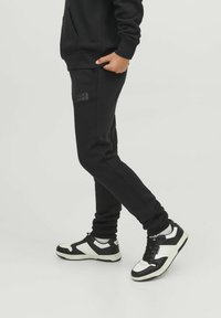 Melni sporta bikses ar slaidu piegriezumu, ar mazu Jack & Jones logotipu, apvienotas ar melnbaltiem masīviem sporta apaviem.