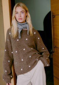 Pull marron avec des motifs de fleurs blanches, texture côtelée et manches larges. Porté avec une écharpe grise. Un pantalon beige est également visible.