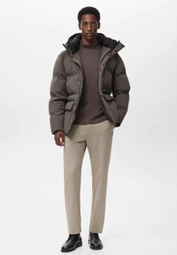 Giubbotto bomber marrone scuro con colletto alto, chiusura con bottoni, due tasche frontali, abbinato a un maglione scuro e pantaloni beige. Scarpe nere.
