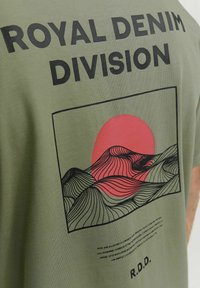 Camiseta de algodón verde oliva con un gráfico negro de olas y un sol rojo, con el texto "ROYAL DENIM DIVISION" arriba y un eslogan debajo.