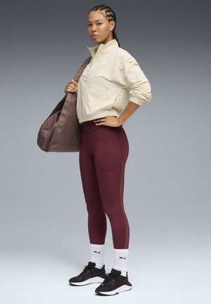 Helle beige Windjacke mit hohem Kragen, bordeauxfarbene Leggings mit seitlichen Einsätzen, schwarze und pinke Sneakers, weiße Socken und eine braune Tasche.