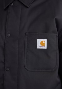 Must mustvalge tööida, mis on valmistatud vastupidavast kangast, kus on terav krae, ees nupud ja vasakul rinnakul tasku koos valge ja oranži logoga.