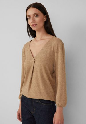 Beige Langarmshirt mit V-Ausschnitt und einer Falten-Detail vorne. Der Stoff hat ein weiches, strukturiertes Aussehen. Kombiniert mit dunkelblauen Jeans.
