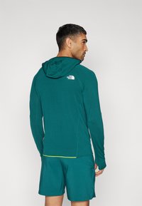 Teal long-sleeve hoodie with a textured fabric and hood, featuring a small white logo on the upper back. Paired with teal shorts.