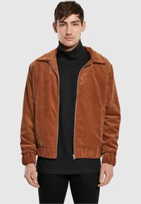 Brown corduroy zip-up jacket with a collar, ribbed cuffs, and a relaxed fit, worn over a black turtleneck. Subtle texture detail visible.